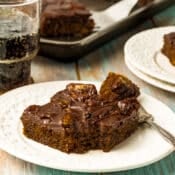 A square piece of chocolate cake with dark frosting on a white plate, next to a fork and a glass of soda, on a light wooden table. Another plate and a baking tray with more cake are in the background.