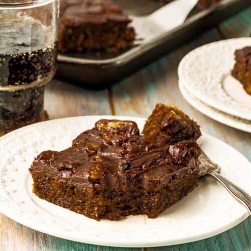A square piece of chocolate cake with dark frosting on a white plate, next to a fork and a glass of soda, on a light wooden table. Another plate and a baking tray with more cake are in the background.