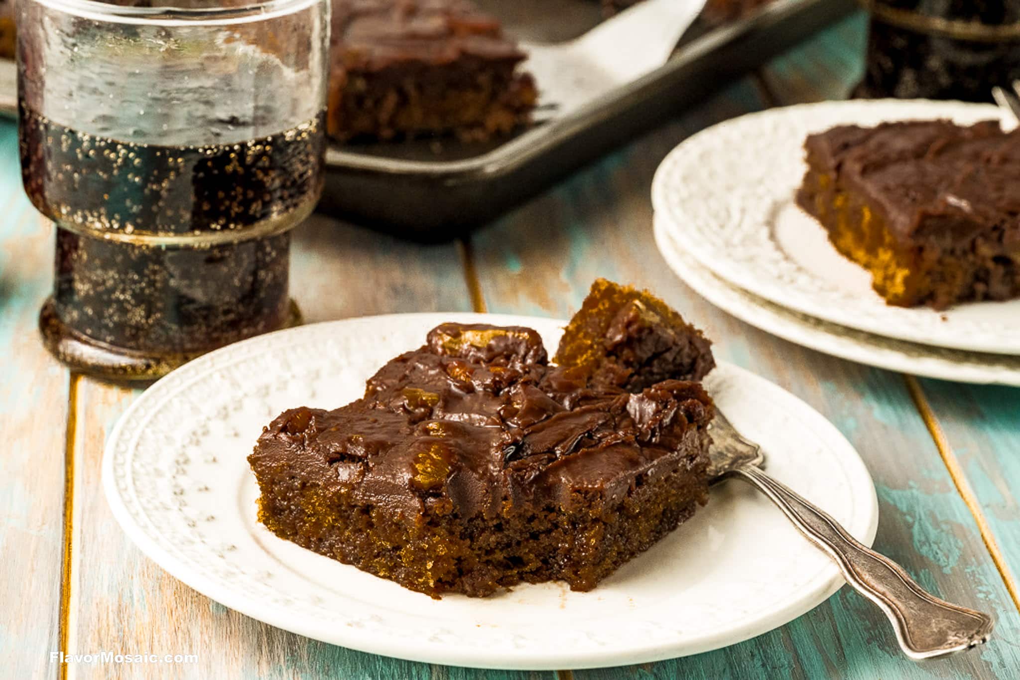 A square piece of chocolate cake with dark frosting on a white plate, next to a fork and a glass of soda, on a light wooden table. Another plate and a baking tray with more cake are in the background.
