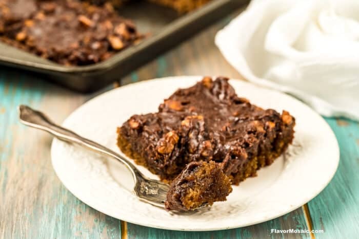 A slice of gooey chocolate dessert with nuts on a white plate, a fork taking a bite, with a baking pan and white napkin in the background on a wooden table.