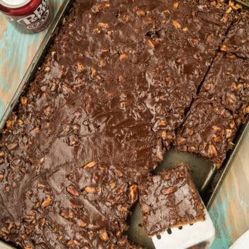 A pan of chocolate sheet cake with nuts, a spatula lifting one square piece, and a can of Dr. Pepper on a wooden surface.