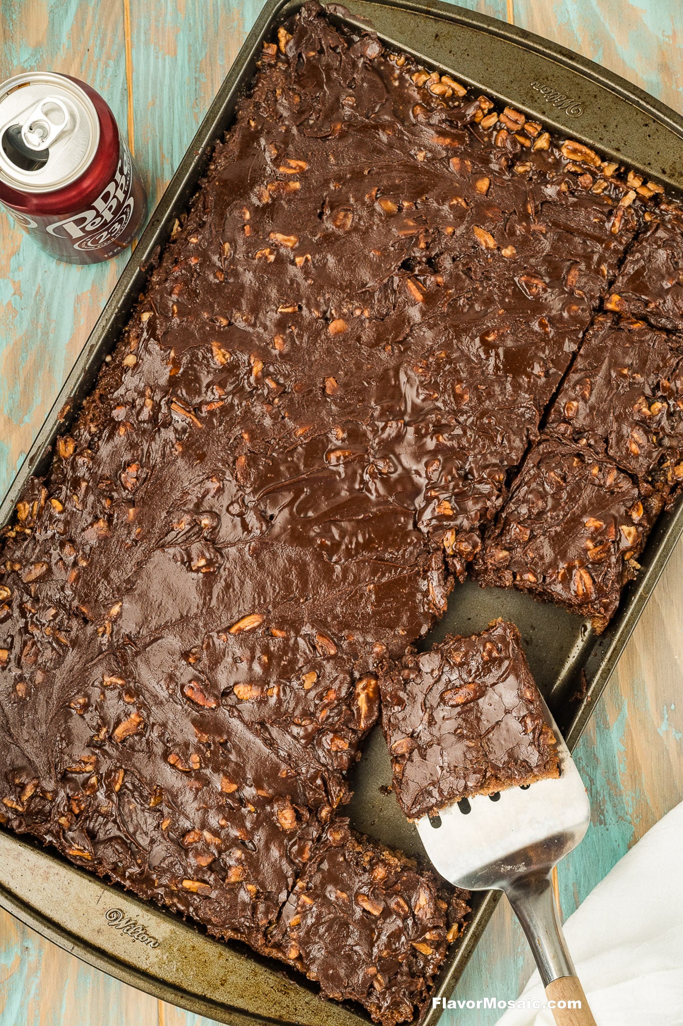 A pan of chocolate sheet cake with nuts, a spatula lifting one square piece, and a can of Dr. Pepper on a wooden surface.
