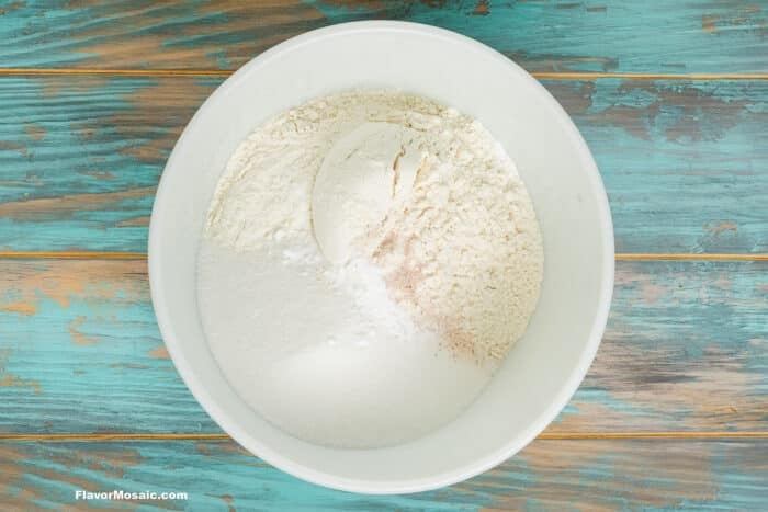 A white bowl on a blue wooden surface contains a mixture of flour, sugar, and baking powder, with the dry ingredients unmixed.