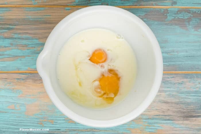 A white mixing bowl contains two cracked eggs and milk, sitting on a distressed blue wooden surface.