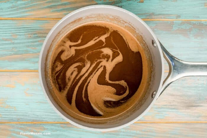 A pot of hot chocolate with swirling foam sits on a rustic, teal and wood-colored surface.