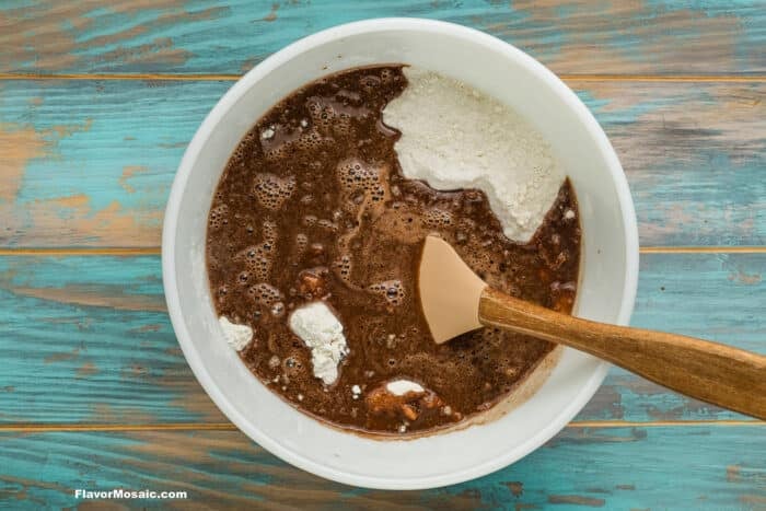 A mixing bowl with brownie batter, some unmixed flour, and a spatula on a blue wooden surface.