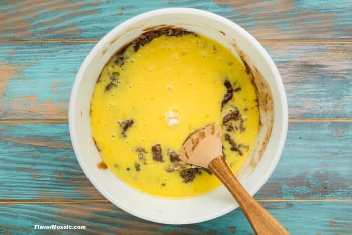 A white bowl with a mixture of beaten eggs and chocolate, partially blended, sits on a blue wooden surface. A wooden spoon rests inside the bowl.