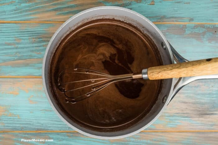 A metal saucepan with chocolate sauce being stirred by a whisk sits on a rustic teal wooden surface.