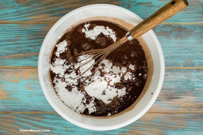 A white bowl with chocolate batter and white flour being mixed with a whisk, on a rustic blue wooden surface.