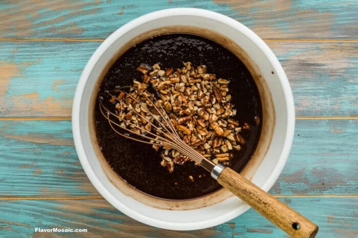 A white bowl with a dark liquid mixture and chopped nuts, being stirred with a whisk on a rustic blue and brown wooden surface.
