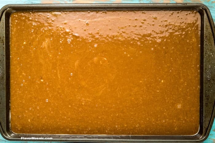A dark baking sheet is filled with an even layer of smooth, glossy brown batter, ready to be baked. The surface is speckled and slightly textured.