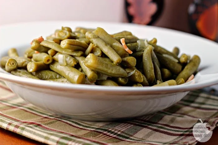 A white bowl filled with cooked green beans sits on a plaid cloth. The green beans appear to be seasoned and garnished with small bits of onion or bacon.