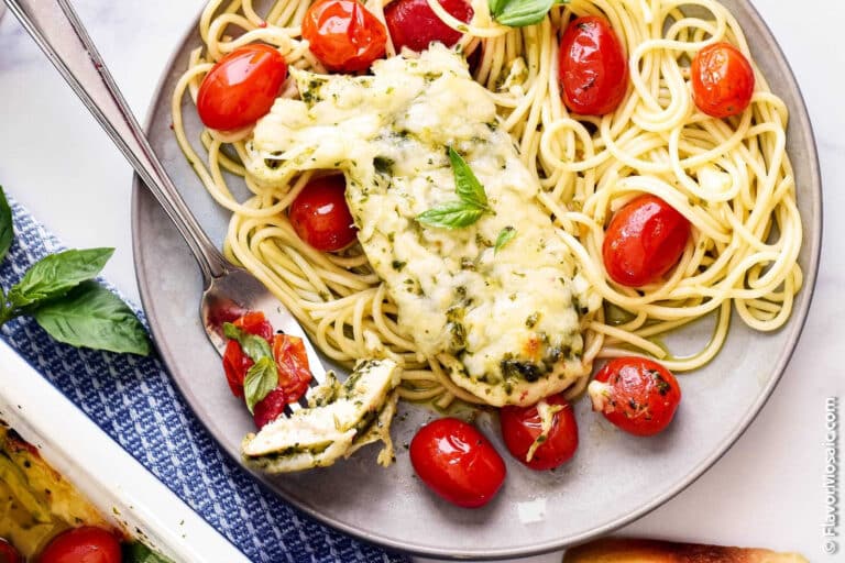 Plate of spaghetti with roasted cherry tomatoes, a baked chicken breast topped with melted cheese and herbs, garnished with fresh basil. A fork holds a piece of chicken.