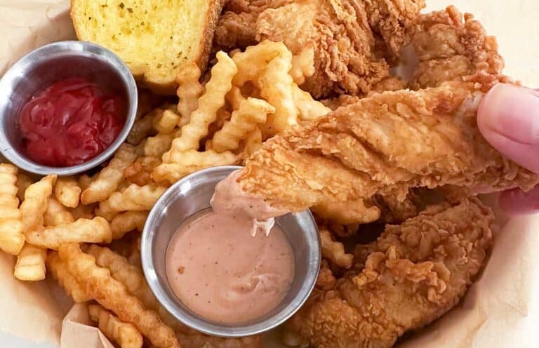 A basket of crinkle-cut fries, fried chicken tenders, a slice of toast, ketchup, and dipping sauce-just like your favorite American Restaurant Copycat Recipes. A hand dips a chicken tender into a creamy sauce, capturing the perfect bite.