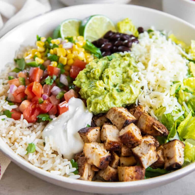 A burrito bowl with grilled chicken, rice, shredded cheese, black beans, corn, pico de gallo, guacamole, lettuce, sour cream, and lime wedges in a white bowl-perfect for anyone craving American Restaurant Copycat Recipes at home.