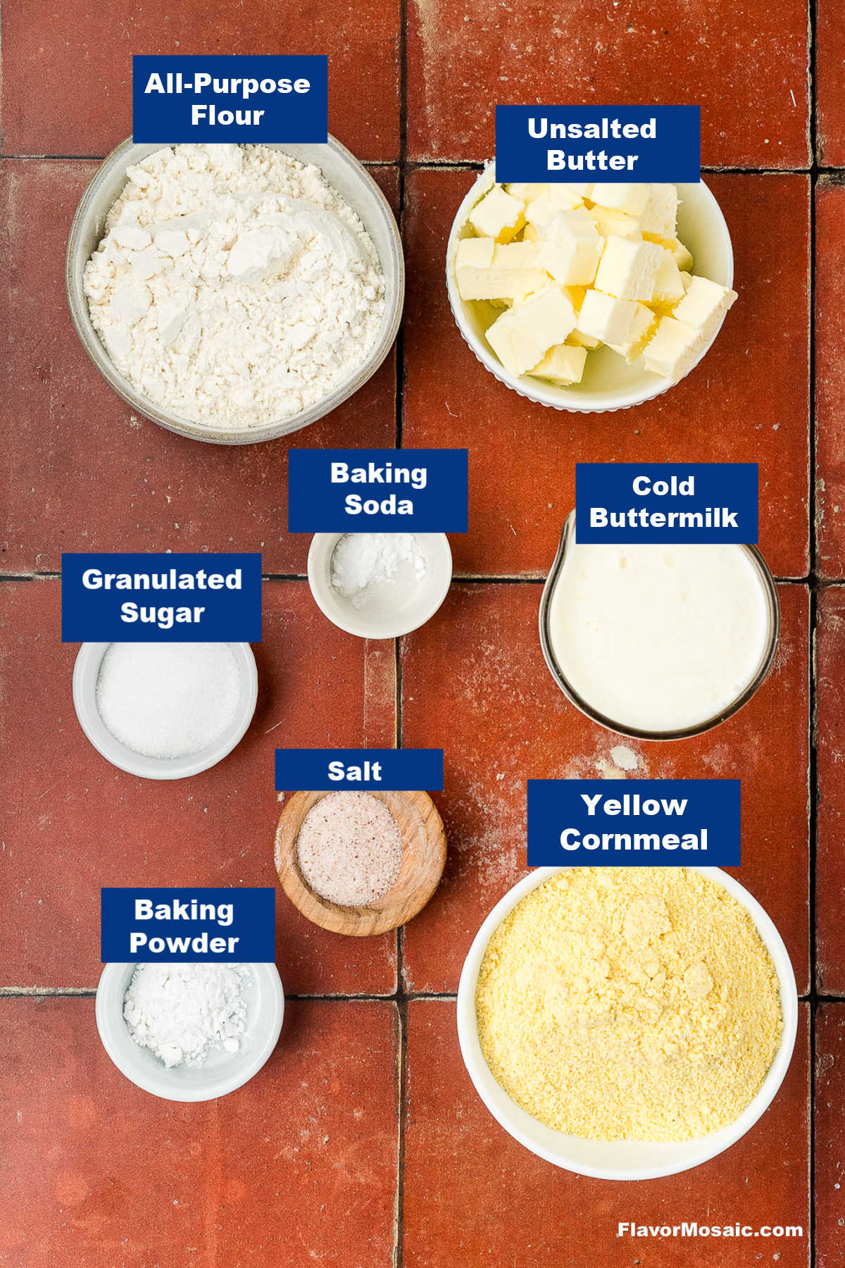 Bowls on a red tile surface hold labeled baking ingredients: all-purpose flour, unsalted butter, cold buttermilk, yellow cornmeal, granulated sugar, baking soda, baking powder, and salt.