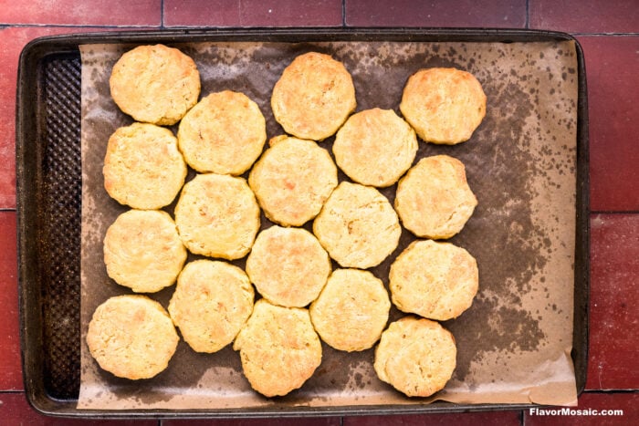 A baking tray with 18 golden brown biscuits arranged closely on parchment paper, set on a red tiled surface.