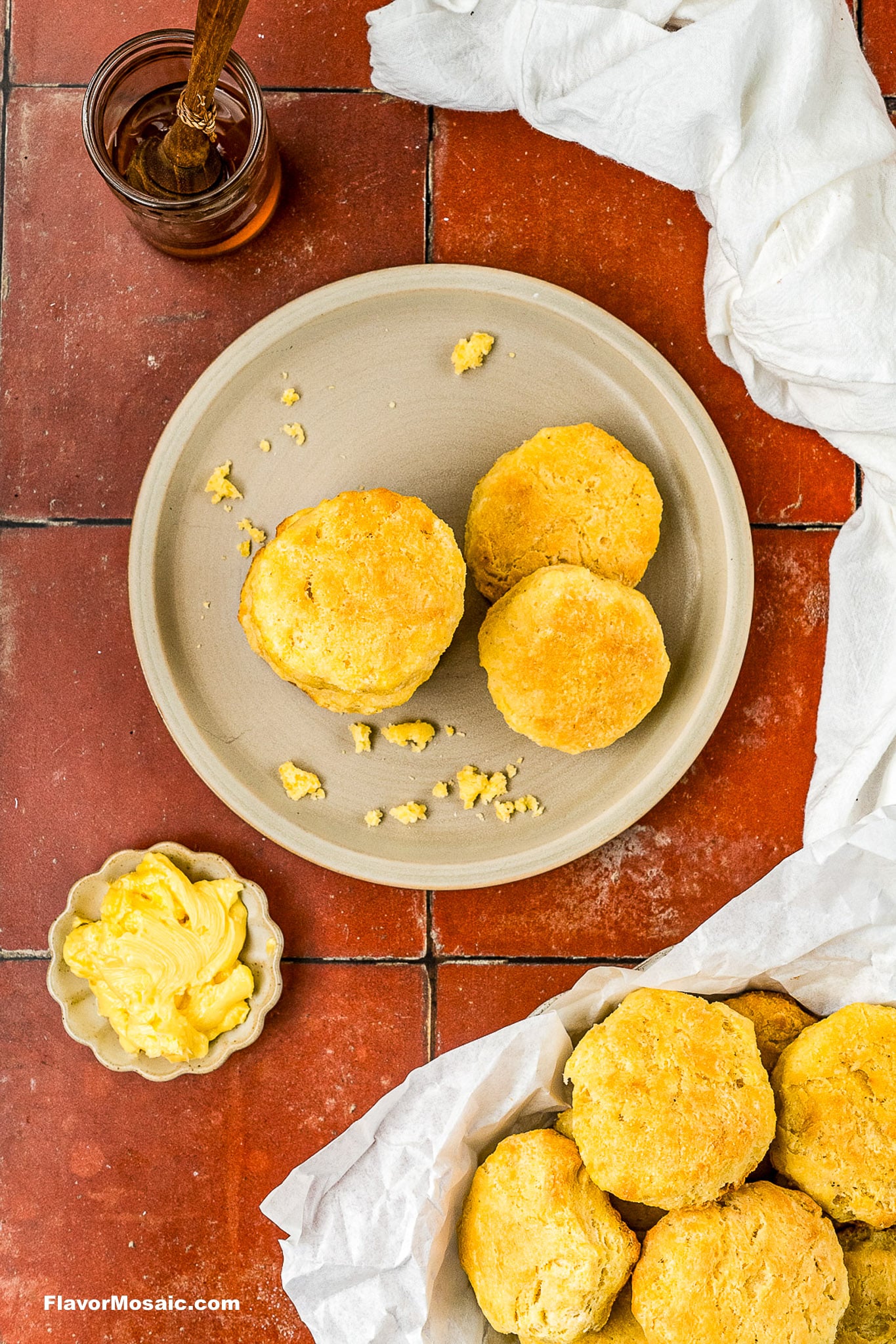 A beige plate with three golden biscuits, surrounded by more biscuits in a basket, a small dish of butter, a jar of honey with a dipper, and a white cloth on a red tile surface.