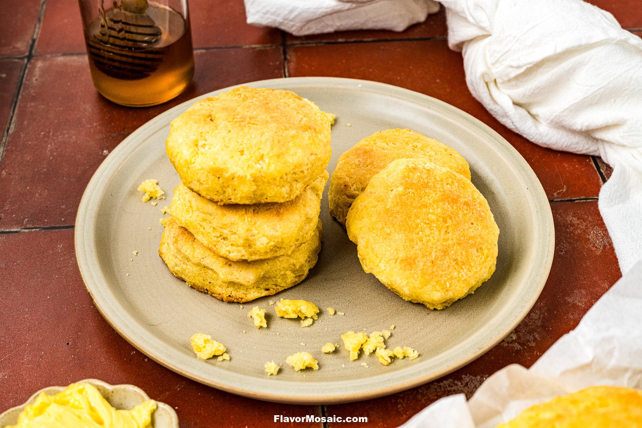 A plate of golden biscuits, some stacked and some lying flat, sits on a red-tiled surface. Crumbs and a pat of butter are nearby, with a glass jar of honey and white cloths in the background.