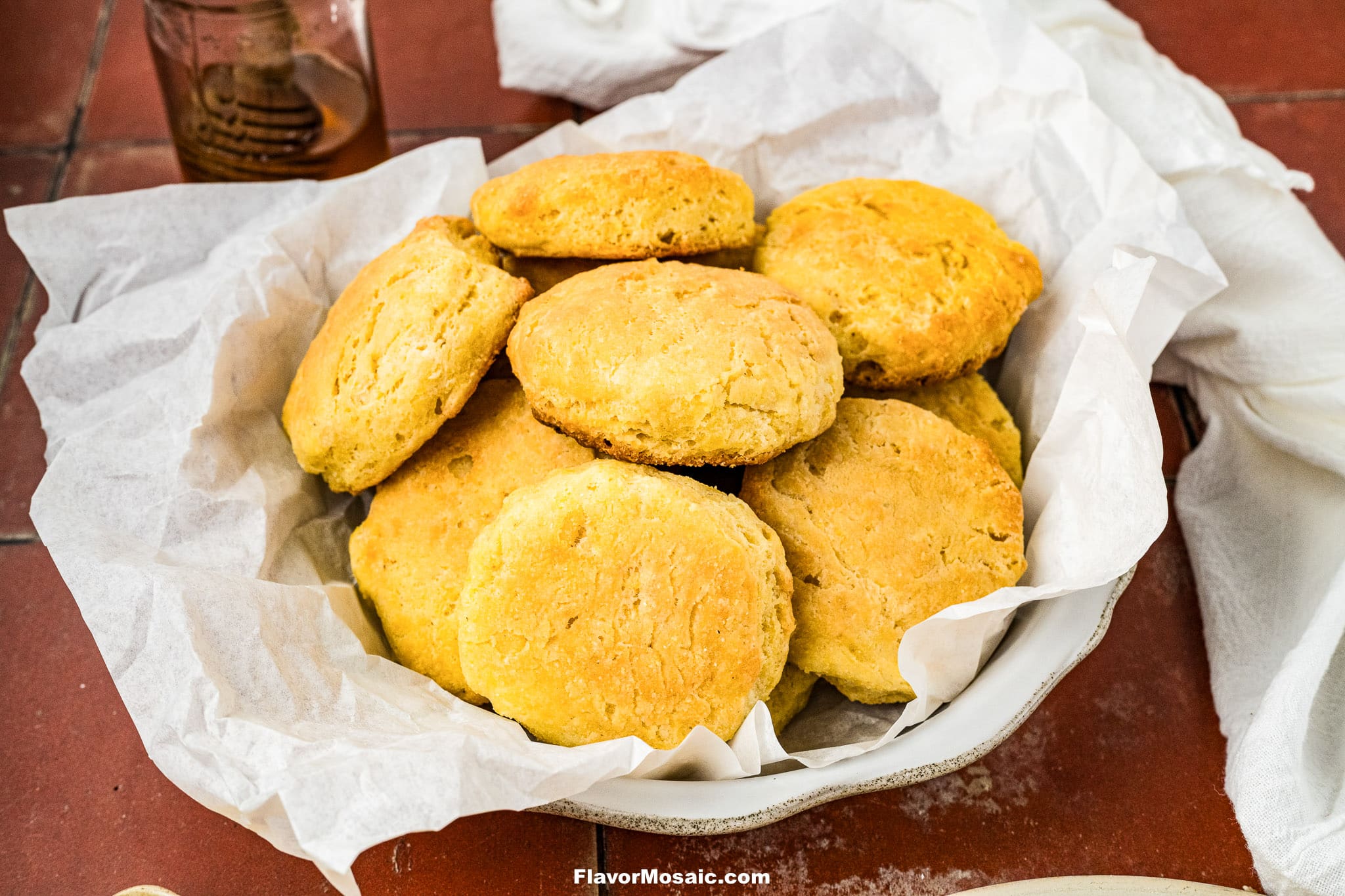 A bowl lined with parchment paper holds golden, freshly baked biscuits. The bowl is on a reddish tabletop, with a jar and white cloth partially visible in the background.
