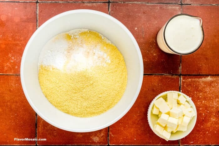 A bowl of cornmeal, salt, and baking powder, a bowl of cubed butter, and a measuring cup of milk sit on a red tiled surface.