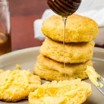 A stack of biscuits sits on a plate. One biscuit is split open with butter melting on top, and honey is being drizzled onto it from a wooden honey dipper.