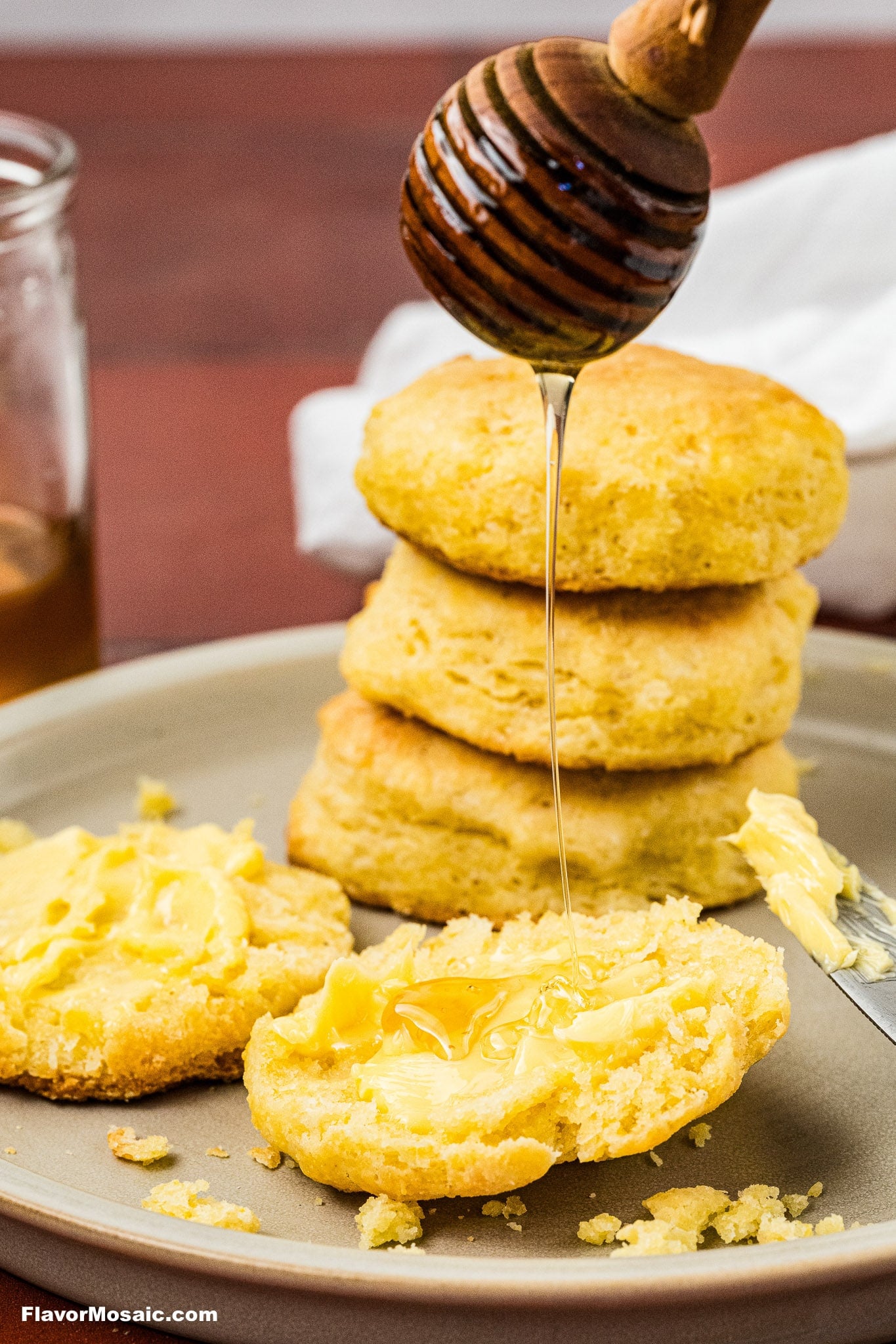 A stack of biscuits sits on a plate. One biscuit is split open with butter melting on top, and honey is being drizzled onto it from a wooden honey dipper.