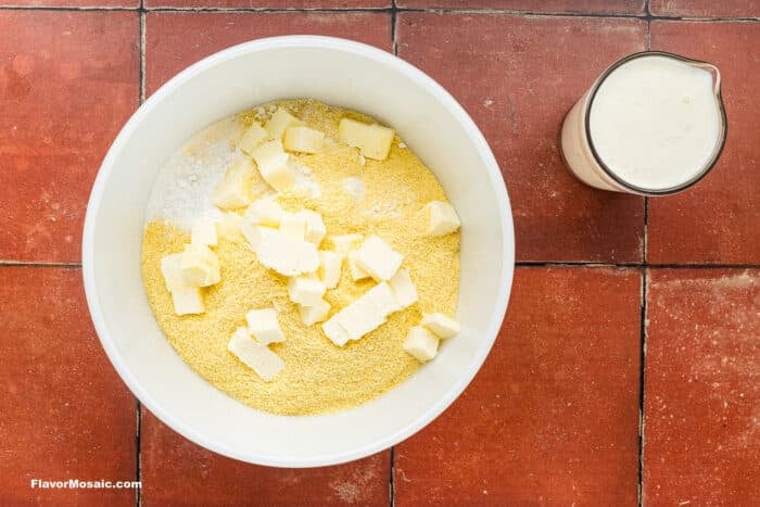 A white bowl with cornmeal, flour, and cubed butter on a red tiled surface, next to a small pitcher of milk.