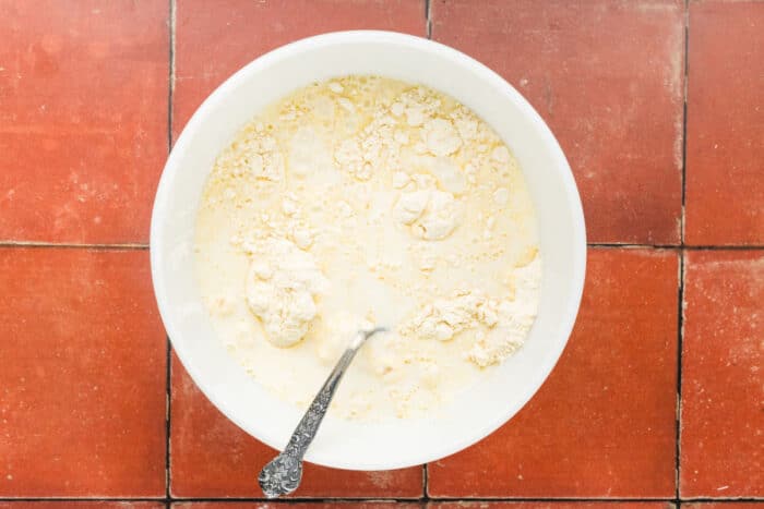 A white bowl filled with flour and liquid, partially mixed, sits on a red tiled surface. A metal spoon rests inside the bowl.
