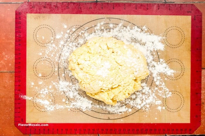 A mound of yellow dough dusted with flour sits on a red-edged silicone baking mat marked with circles and guides, atop a tiled surface.
