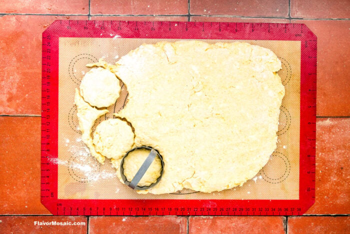 Biscuit dough rolled out on a red silicone baking mat, with round biscuit shapes being cut out using a metal cutter. The surface is dusted with flour.