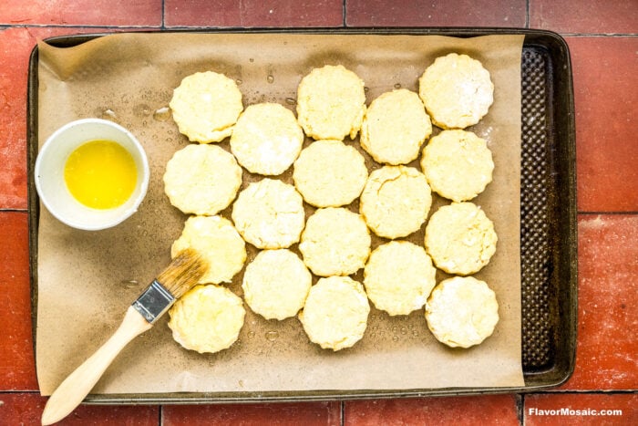 A baking sheet lined with parchment paper holds unbaked biscuit dough rounds. A bowl of melted butter and a pastry brush sit beside the biscuits, ready for brushing. The baking sheet rests on a red tiled surface.