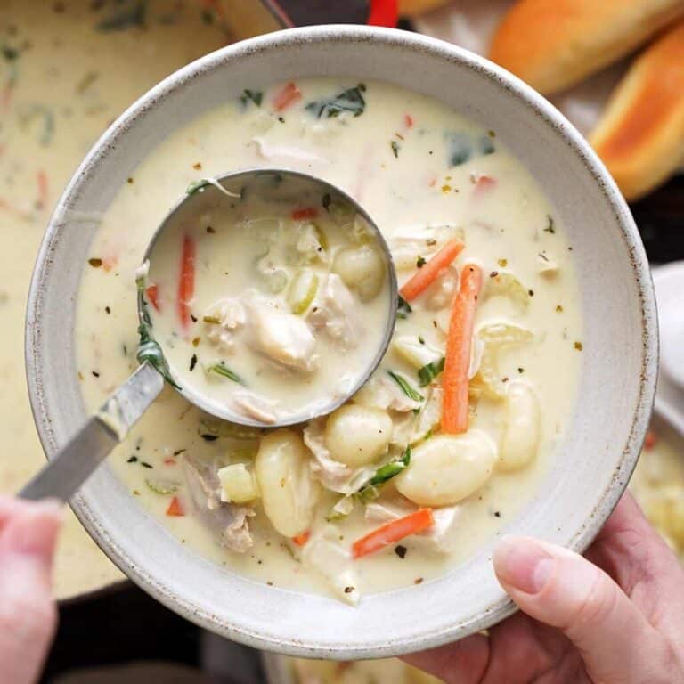 A hand holds a bowl of creamy chicken gnocchi soup with carrots, celery, and spinach while a ladle pours more soup into the bowl. Breadsticks are visible in the background, evoking the comforting flavors found in popular American Restaurant Copycat Recipes.
