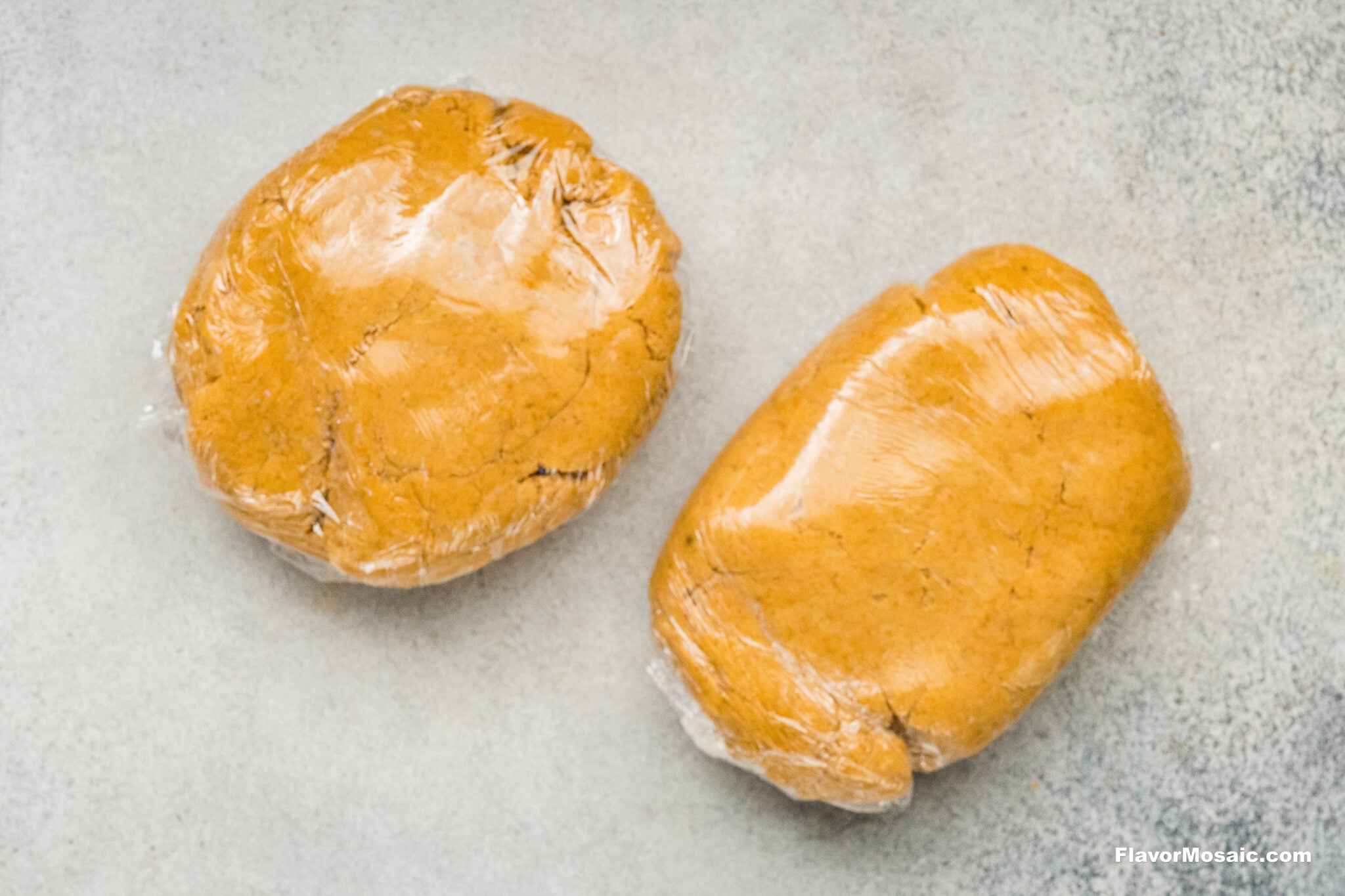 Two balls of pumpkin spice sugar cookie dough wrapped in plastic wrap on a light countertop.