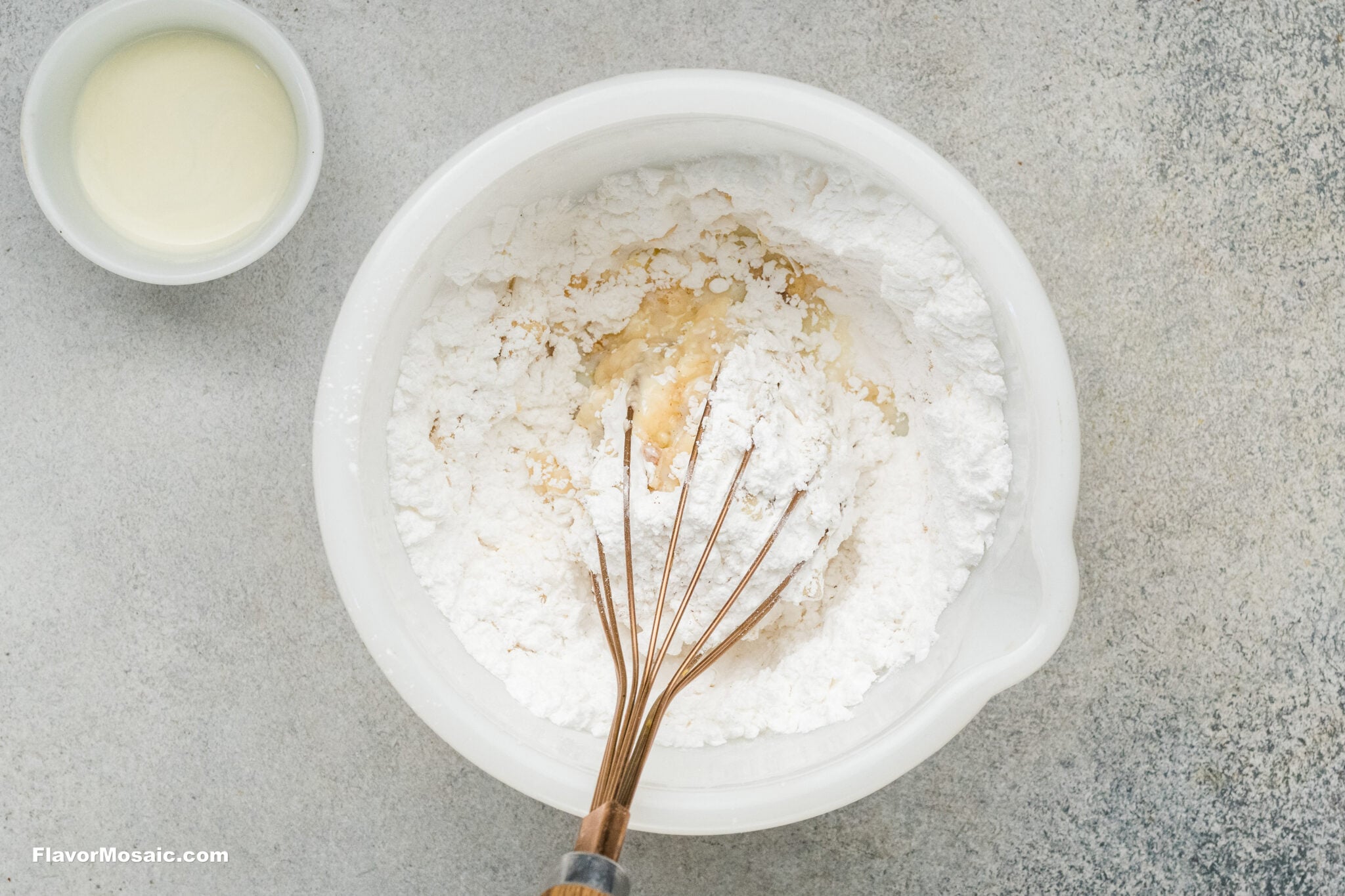 A wire whisk mixing flour and liquid in a white bowl for pumpkin spice sugar cookies, with a small dish of liquid to the side.