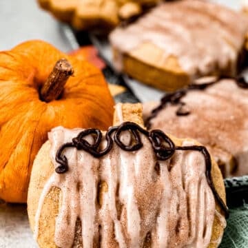 Pumpkin-shaped cookies with cinnamon glaze and chocolate drizzle, next to a small decorative pumpkin.