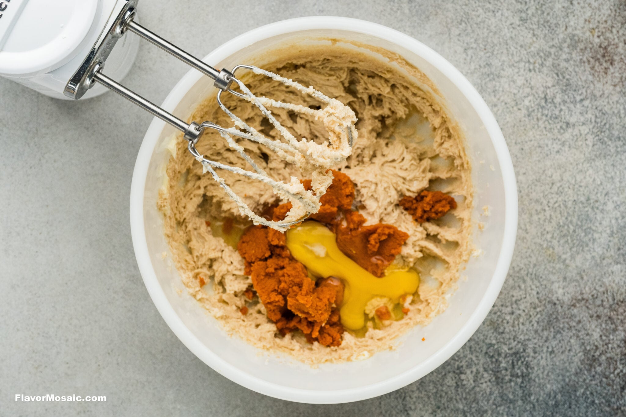 An electric hand mixer with beaters sits over a white mixing bowl containing creamed butter, a cracked egg, and scoops of pumpkin puree for pumpkin spice sugar cookies.