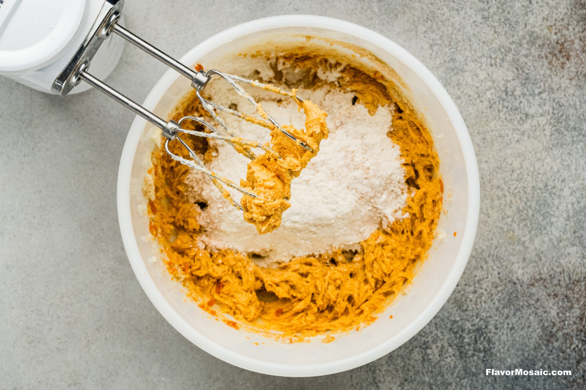 Electric hand mixer blending flour into orange pumpkin spice cookie dough in a white mixing bowl.