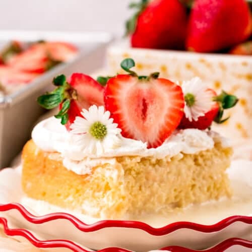 A slice of Tres Leches cake topped with whipped cream, strawberry halves, and small white flowers on a scalloped plate.
