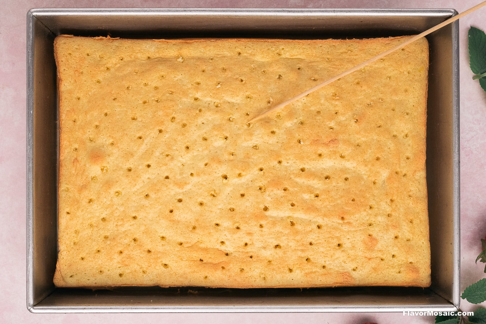 A rectangular Tres Leches cake in a metal baking pan with a wooden toothpick inserted into the top to check for doneness.