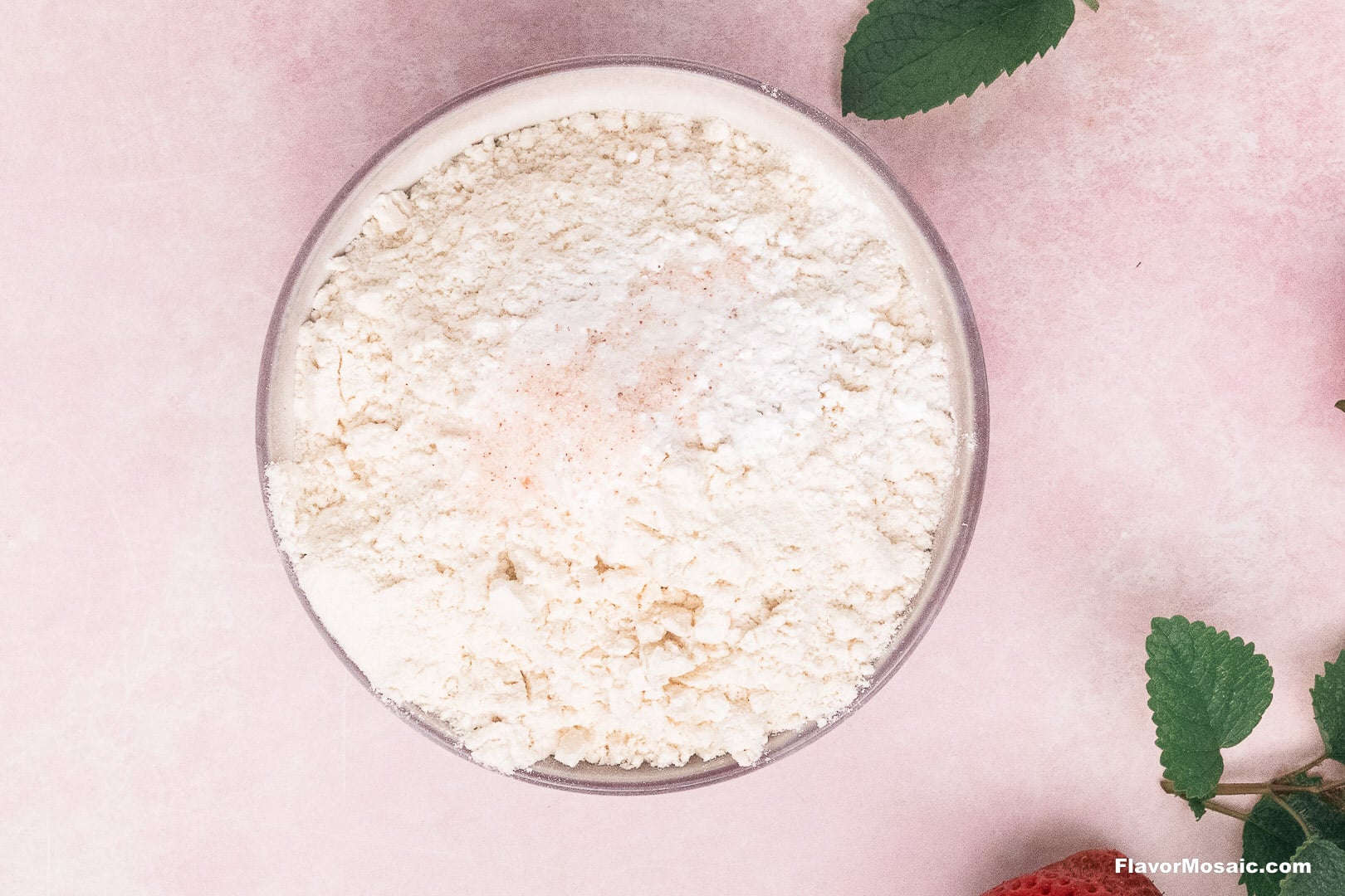 A glass bowl filled with white flour and a sprinkle of pink salt for a Tres Leches Cake recipe, viewed from above next to green leaves and a strawberry.