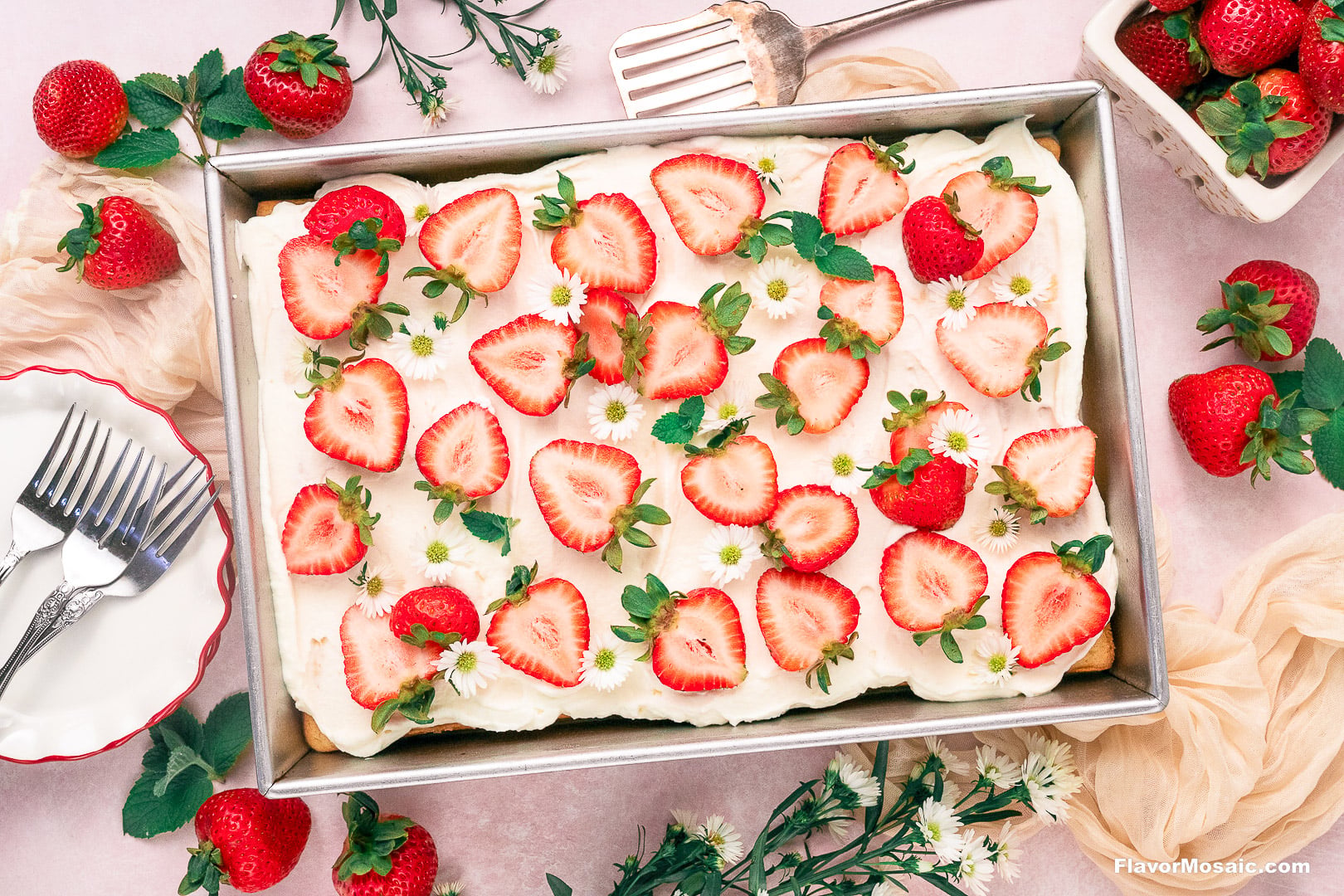 A strawberry tres leches sheet cake topped with white frosting, strawberry slices, and small white flowers, with extra strawberries and forks on the side.