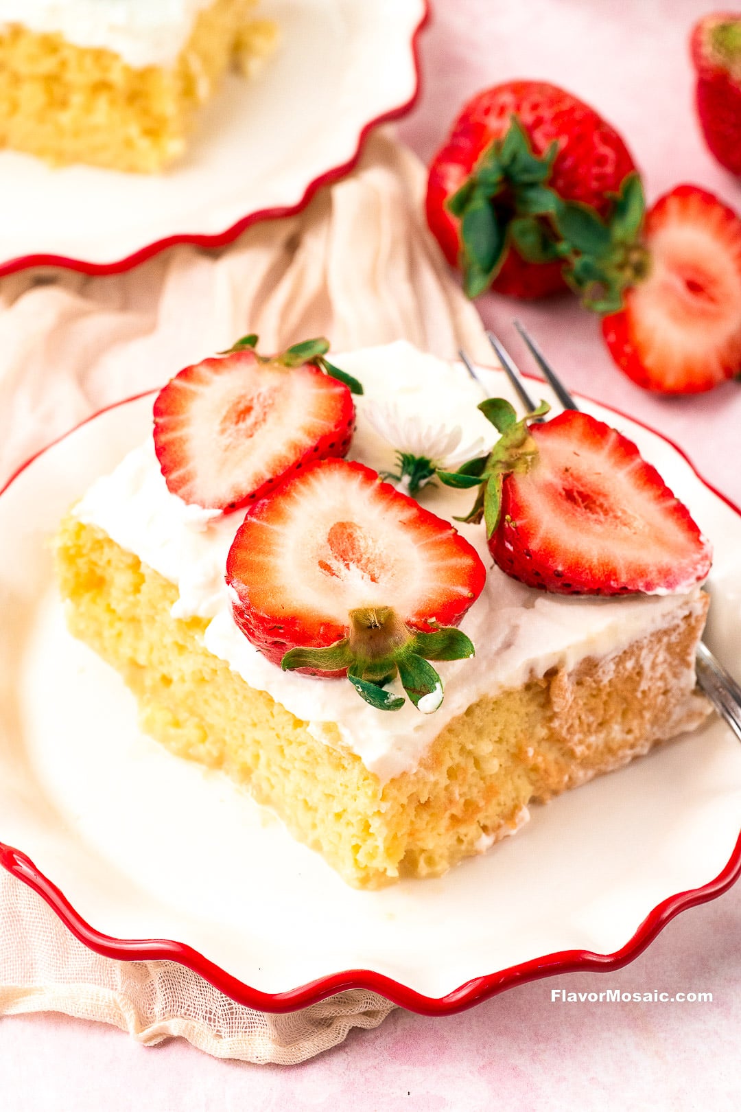 A square slice of yellow tres leches cake topped with white frosting and fresh strawberry slices on a white plate with a red scalloped rim.
