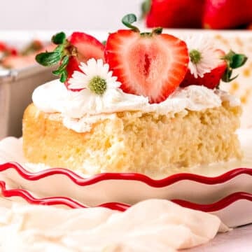 A square slice of strawberry Tres Leches cake topped with whipped cream and small white flowers on a scalloped-edge plate.