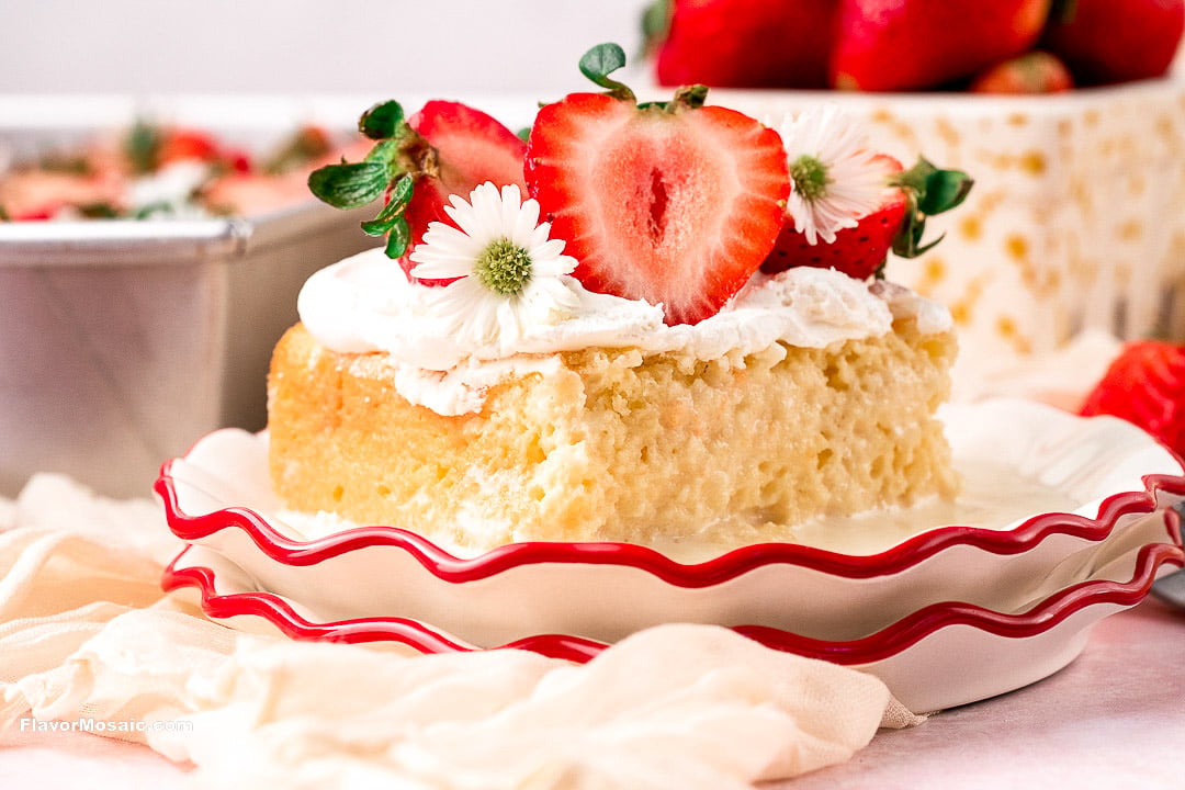 A square slice of strawberry Tres Leches cake topped with whipped cream and small white flowers on a scalloped-edge plate.